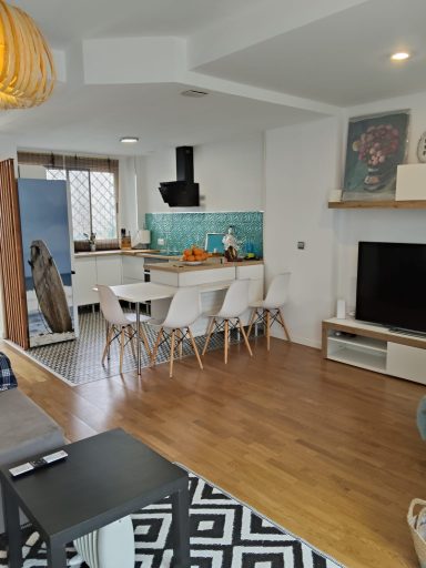 Salón moderno con cocina, mesa blanca, sillas y decoración en tonos azules y madera.