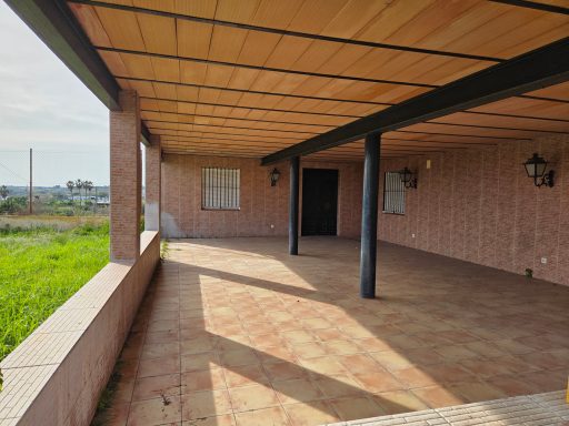 Porche amplio con suelo de cerámica y columnas, rodeado de vegetación.