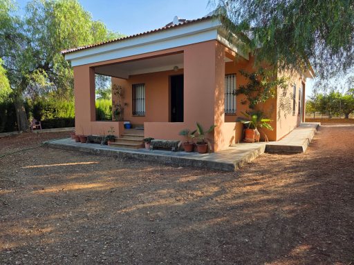 Casa de campo de color rosa con porche y jardín, rodeada de árboles y terreno arenoso.