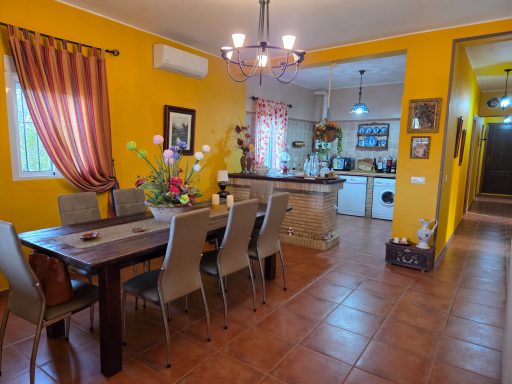 Comedor con mesa larga y sillas, decoración color amarilla y cocina al fondo.
